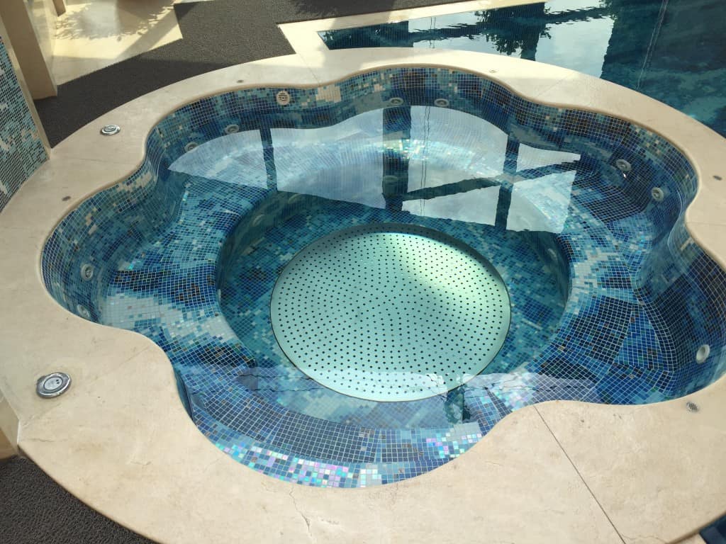 A modern jacuzzi with blue mosaic tiles and a perforated bottom, embedded in a beige floor.