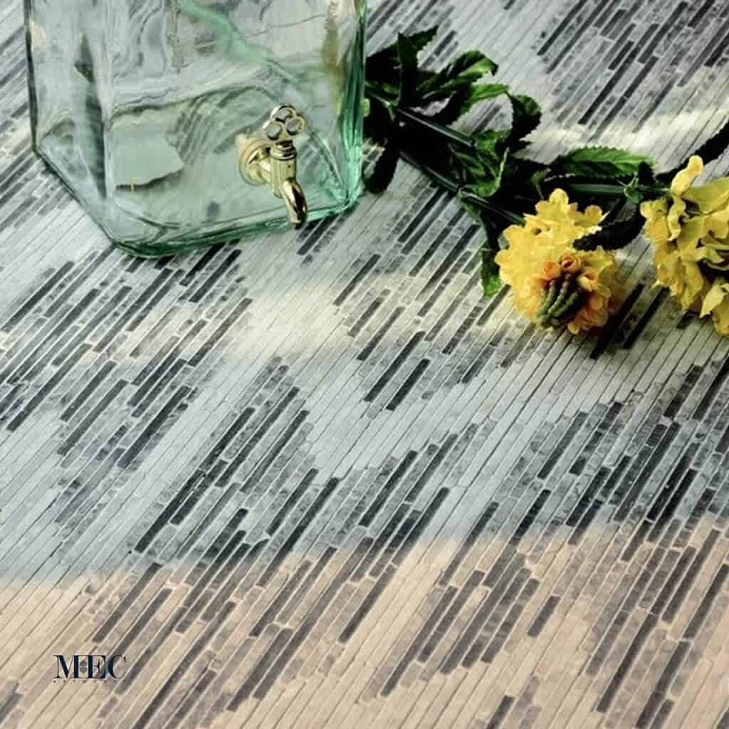 Shevren, an Ikat style marble mosaic floor design made with handchopped gray and white marble stone strips shown with yellow faux flowers and a glass pitcher
