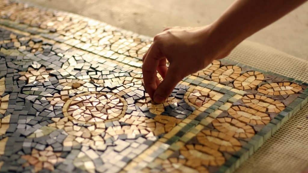 Mosaic floor design ideas blog banner image depicting a craftsman manually placing a marble mosaic piece into a marble rug by MEC. Golden hour light. Close up.