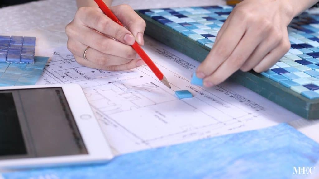 How to choose a pool mosaic design and color? Behind the scenes of color study and decision making process. Close up of hand holding a pencil and a Murano glass tile. Building plan, iPad and PIXL grid in the background.