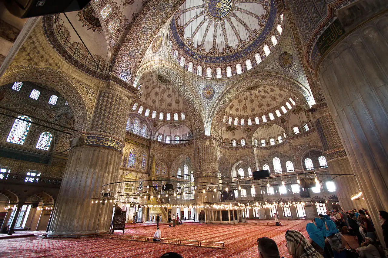 Grand interior view of the Blue Mosque in Istanbul, featuring ornate mosaic tilework, soaring domes, and arched windows.