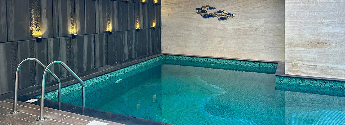 Indoor swimming pool with shimmering turquoise mosaic tiles, modern lighting, and stainless steel ladder.