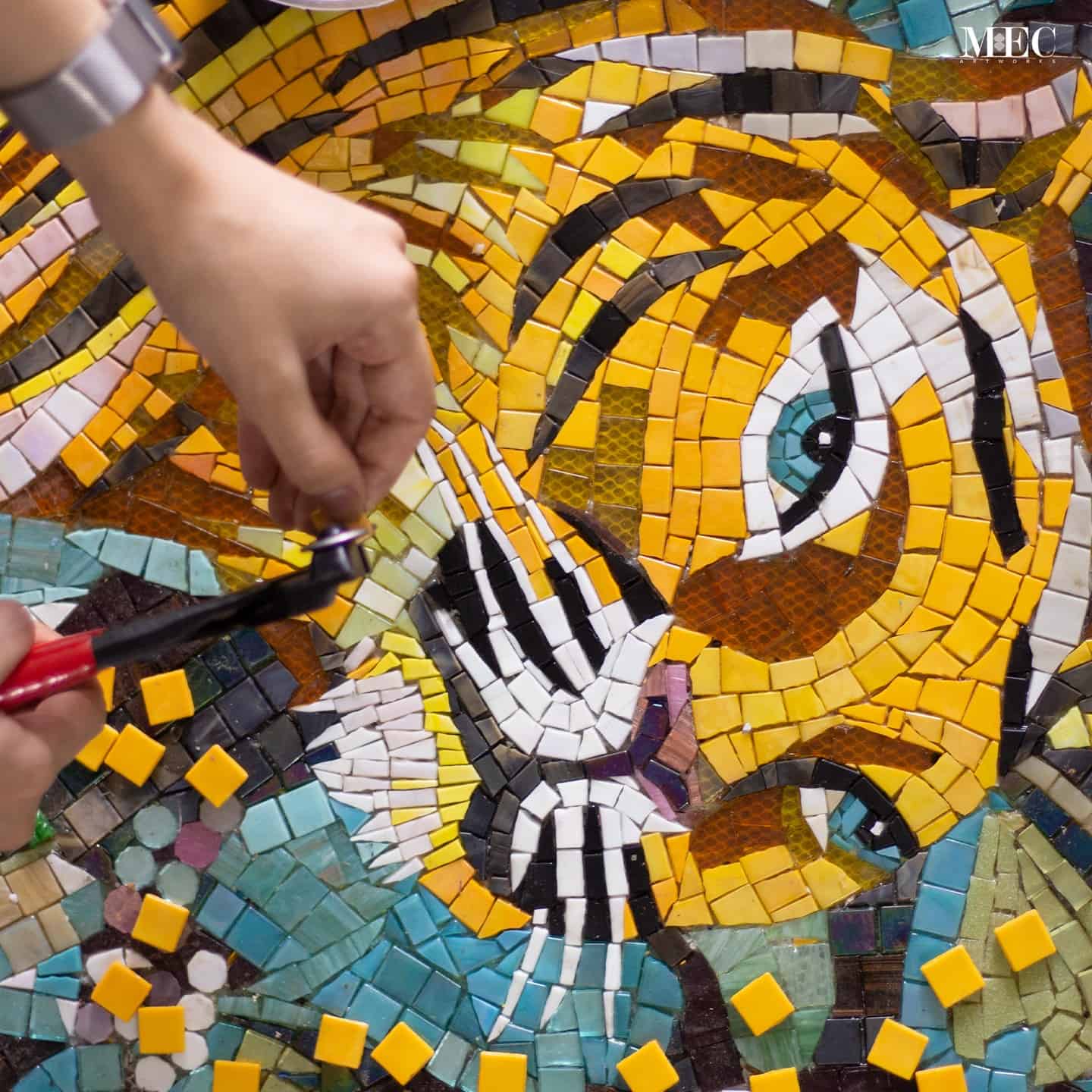 A close-up view of a mosaic artwork of a tiger's face, made of colorful tiles placed by hand with tweezers