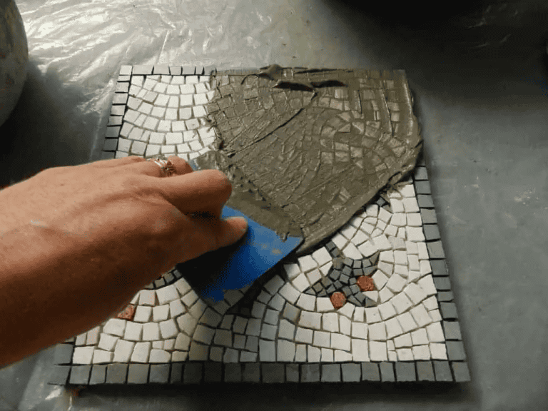 Hand applying grout over small mosaic tiles on square board using blue tool for finishing.