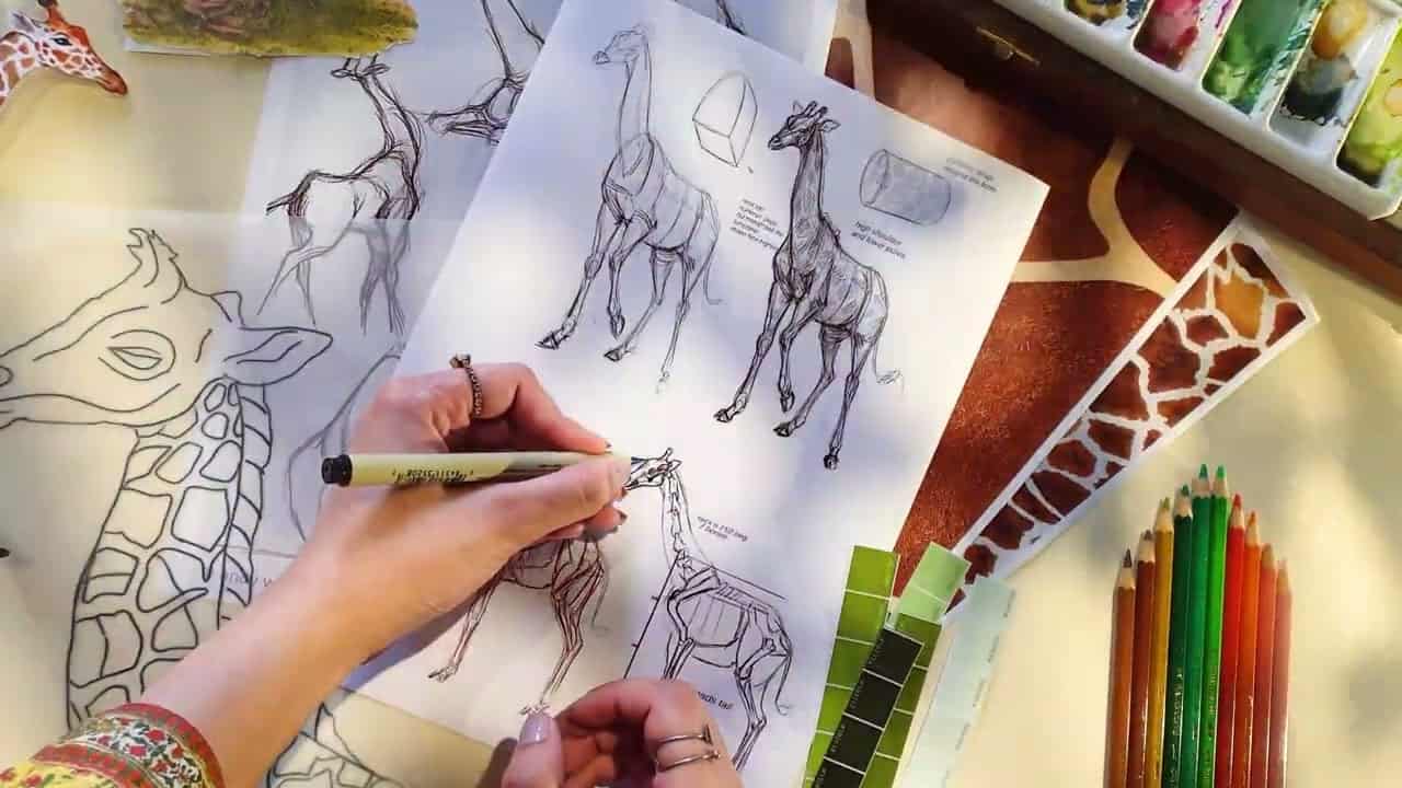 Artist sketching giraffes with reference sheets, color swatches, watercolor palette, and colored pencils nearby.