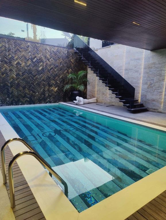 Modern swimming pool with striped blue mosaic tile flooring, metal handrails, and minimalist sculpture near stairs.