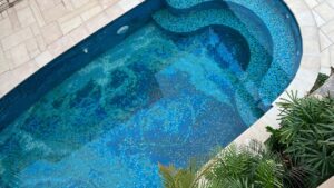 Curved mosaic swimming pool with varying shades of blue tiles and tropical greenery around the edge.