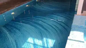 Indoor swimming pool with shimmering blue mosaic tiles, soft lighting, and subtle wave-like tile patterns.