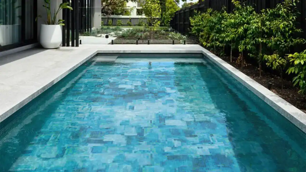 Modern swimming pool with pixelated blue mosaic tiles surrounded by gray paving and lush greenery.