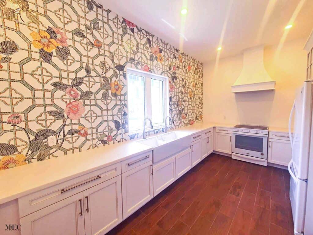 Bright kitchen with white cabinetry, wood flooring, and a vibrant floral mosaic backsplash featuring green geometric patterns.