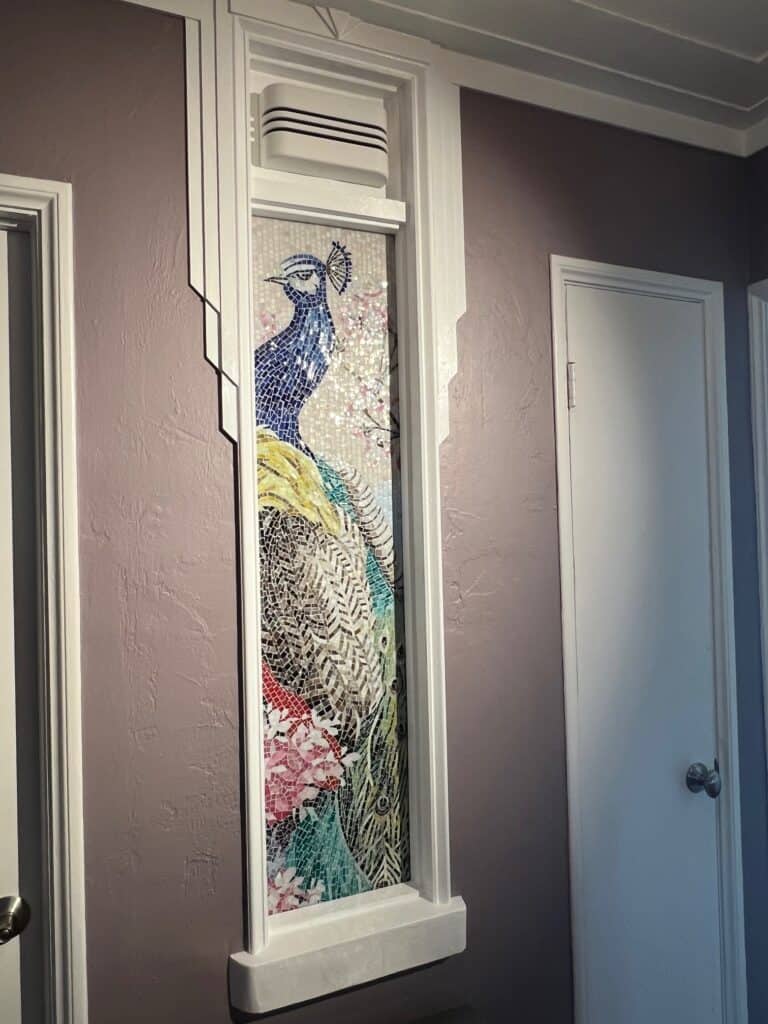 Vertical wall mosaic of a colorful peacock framed in white molding, set between two doors.
