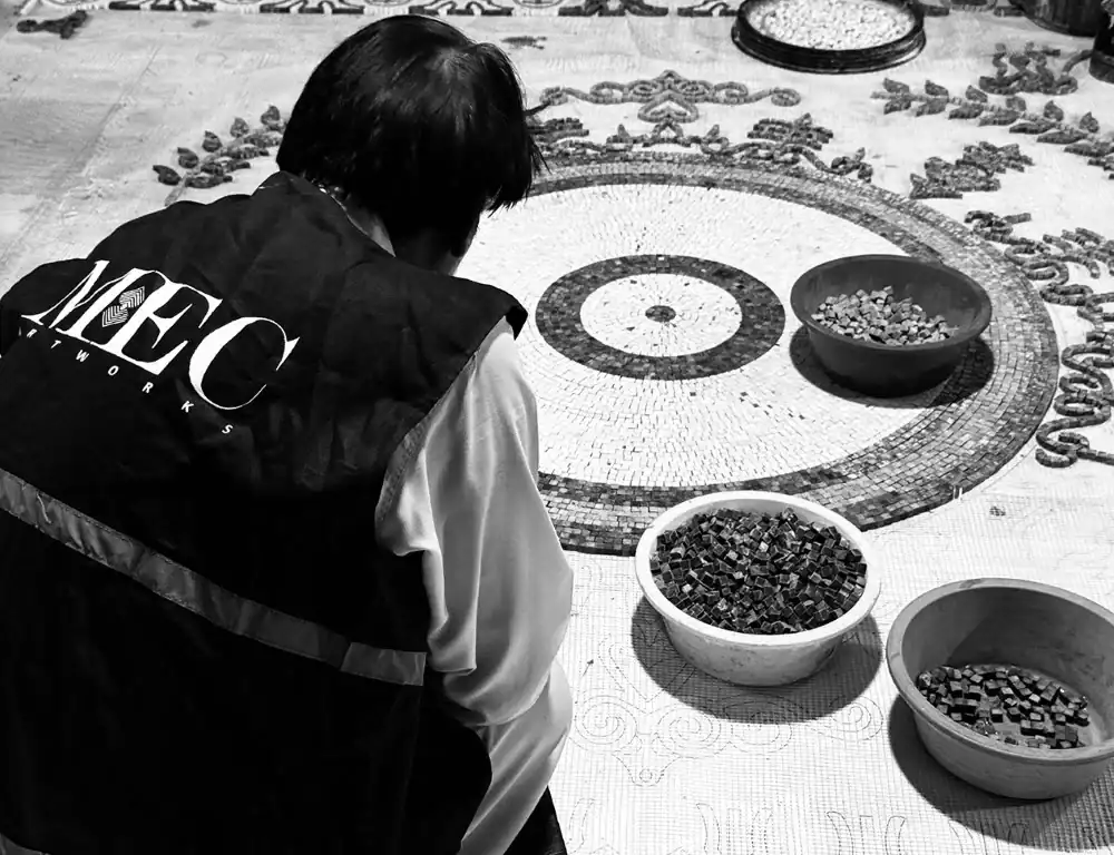 MEC artisan arranging mosaic tiles on circular pattern floor, surrounded by bowls of glass pieces.
