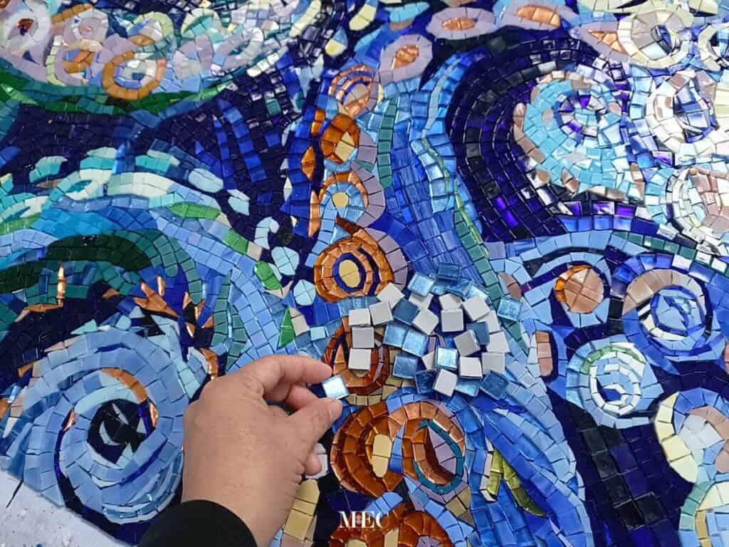 Hand placing small mosaic tiles on swirling blue and orange octopus-themed glass mosaic artwork.