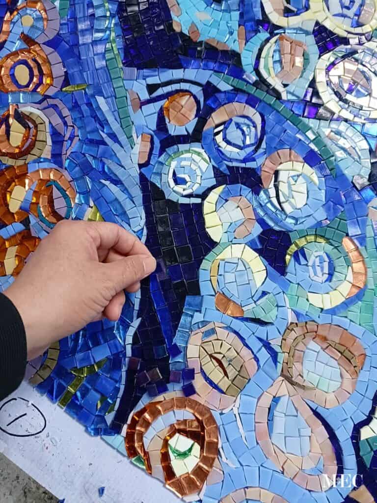 Hand placing mosaic tiles in a vibrant octopus design with swirling blue and copper patterns.