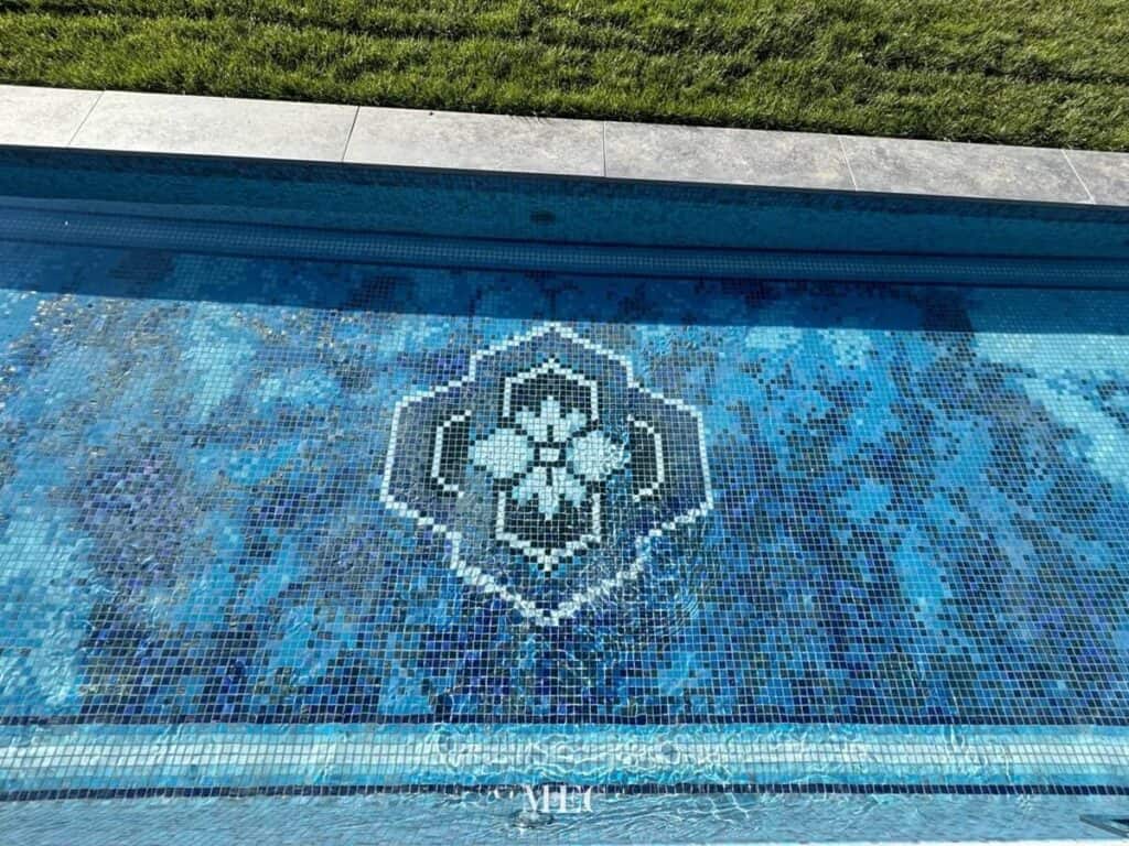 Blue glass mosaic swimming pool with a central white floral medallion and gradient background.