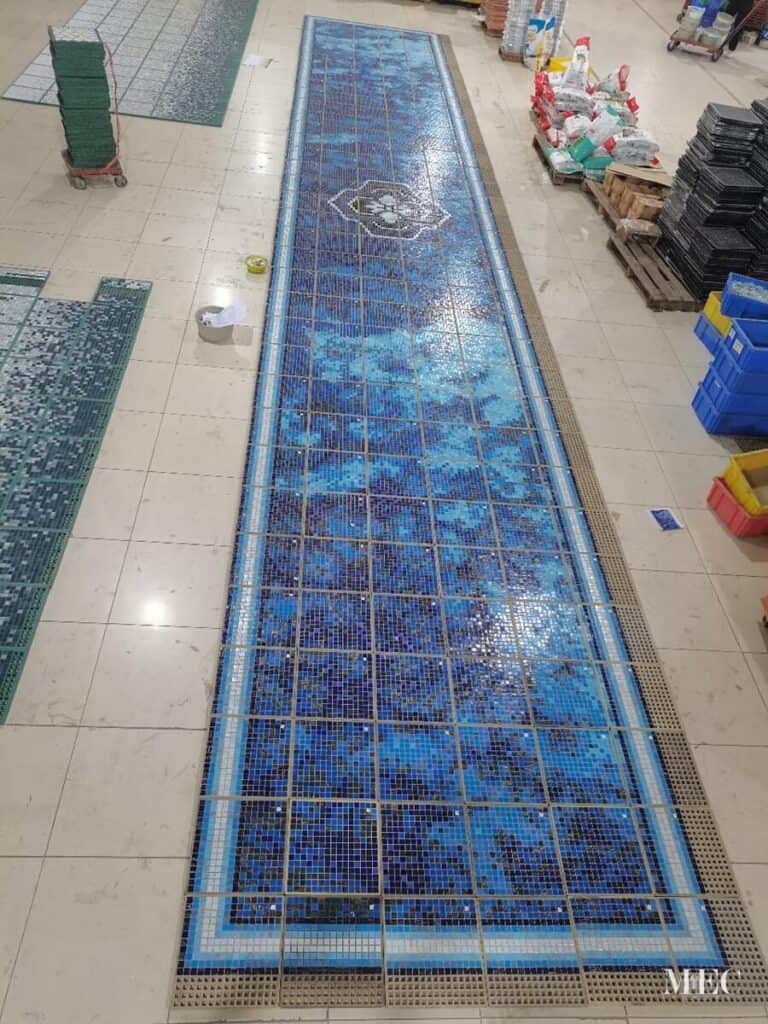 Long blue mosaic pool tile layout with central medallion motif in workshop setting for installation.