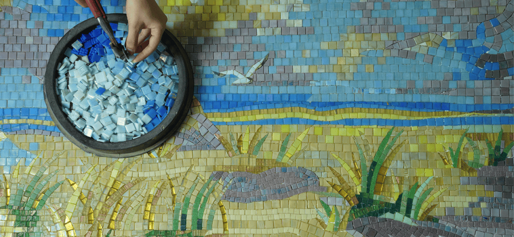 Hand placing blue mosaic tiles into a seaside landscape artwork featuring beach, sky, and birds.