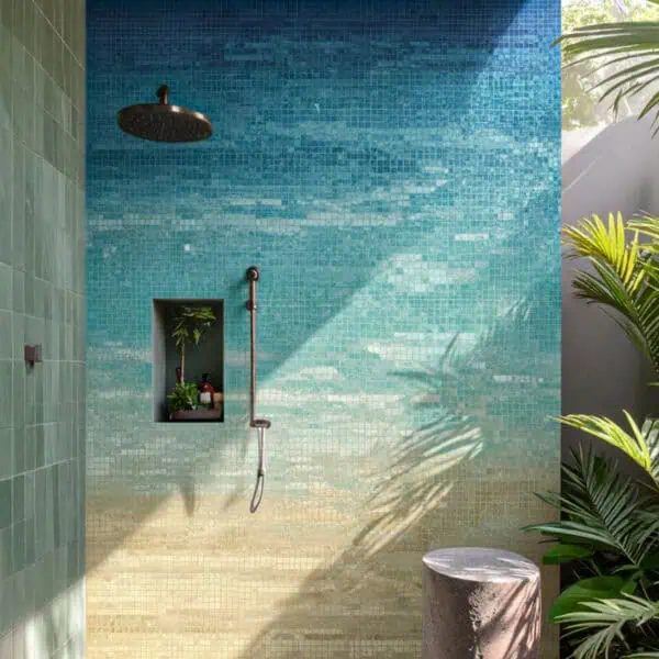 Coastal Horizon Mosaic Shower Outdoor shower with a beach-inspired glass mosaic wall featuring a gradient from sandy beige to ocean blue, complemented by bronze fixtures, plants, and a wooden ceiling.
