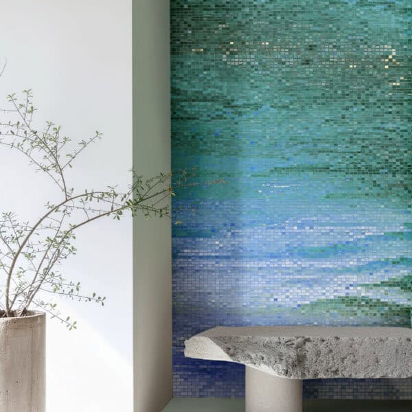 Modern interior featuring a glass mosaic wall with a blue-to-turquoise gradient resembling calm water, paired with a sculptural stone bench and minimalist decor.