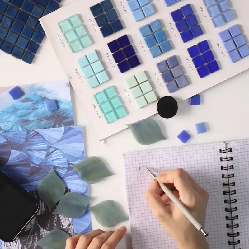 Hand designing tile layout with blue mosaic samples, color swatches, leaves, and a gridded sketchbook.