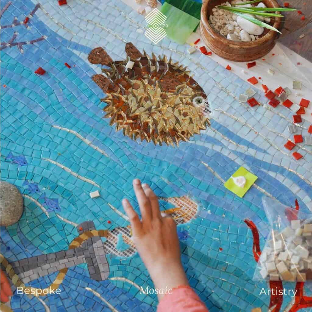 A hand meticulously working on an aquatic mosaic art with blue glass tiles along with a pufferfish above.