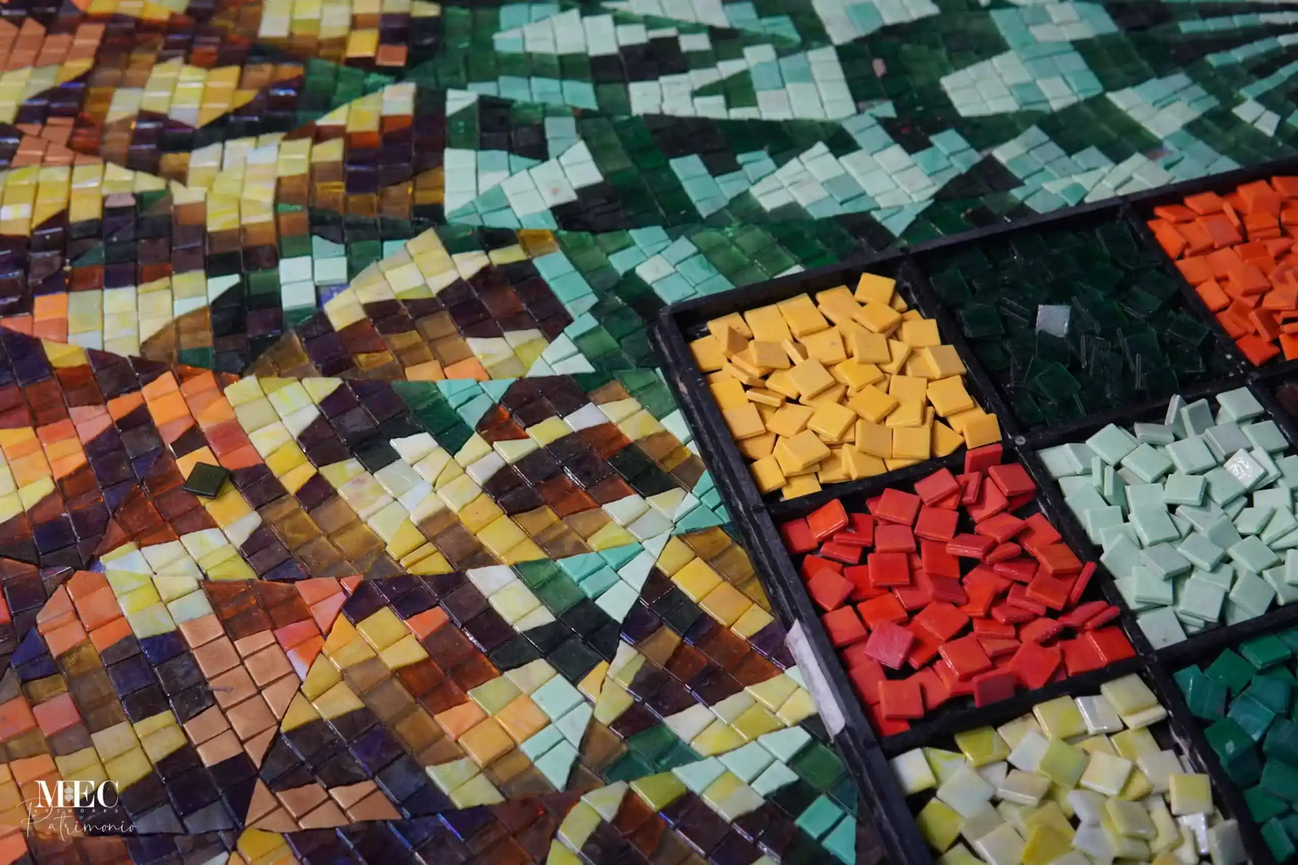 A multi-colored glass mosaic artwork in progress, showcasing sorted trays of vibrant yellow, red, orange, light green, and dark green square tiles next to an assembled patterned layout.