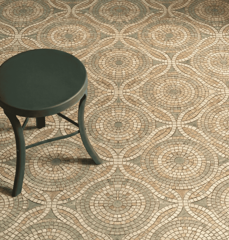 Interlocking orbital circles on stone mosaic floor and curved geometric weaving in earthy tan, beige, and subtle sage green tiles in midst of a minimalist dark green stool.