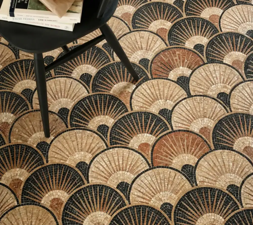 A premium floor layout featuring elegant geometric mosaic tile designs for modern floors, showing an intricate repeating art deco fan pattern hand-assembled in gold, terracotta, beige, and charcoal stone beneath followed by a black chair.