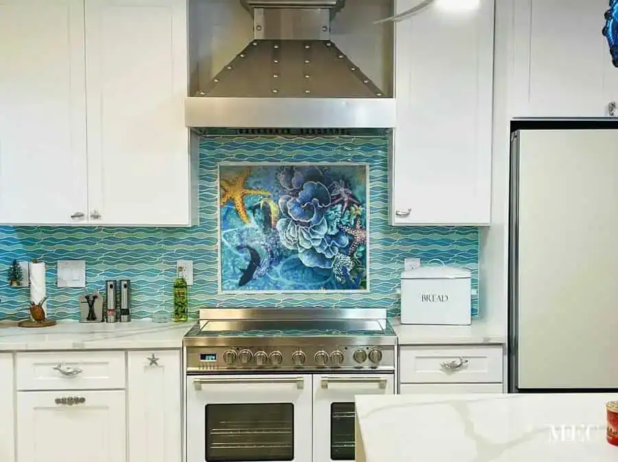 An intricate marine life mosaic work displayed in a kitchen just above a silver stove, showcasing yellow starfish, fish and marine life in tones of blue