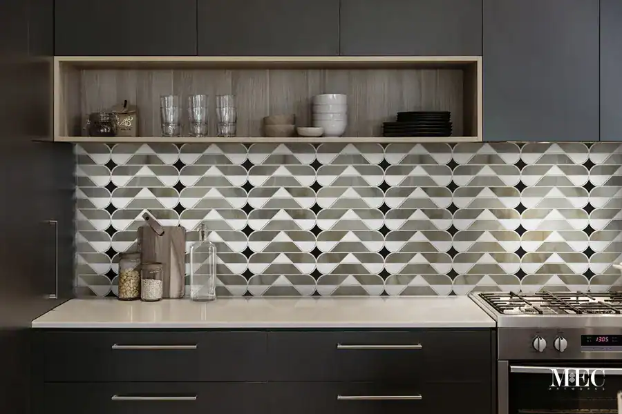 A modern kitchen featuring a geometric mosaic tile kitchen backsplash created through water jet technique with a repeating chevron and curved diamond pattern in grey, white, and black tones over a neutral countertop and dark cabinetry.