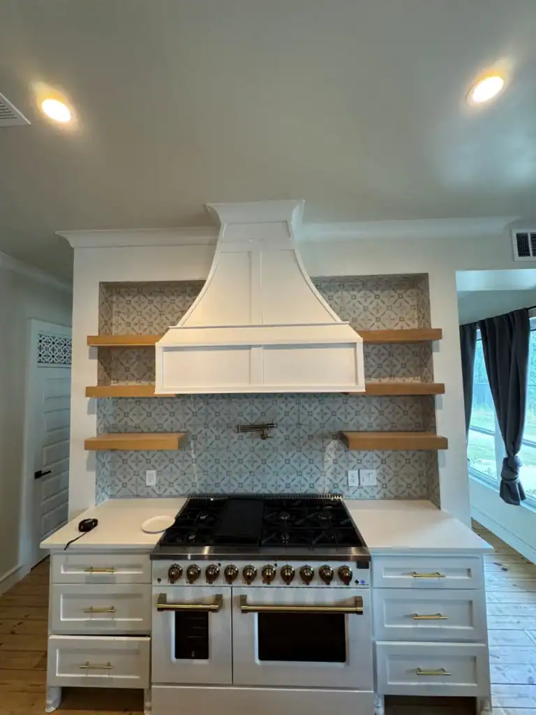 A Frontal view of a kitchen featuring a white gas range stove with brass controls, a white range hood, grey and white geometric patterned tile backsplash, and floating natural wood shelves on either side.