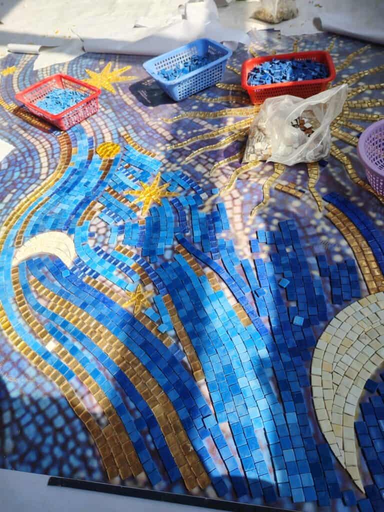 An in-progress mosaic surrounded by small red and blue baskets containing sorted blue glass tiles, with a bag of white tiles nearby.