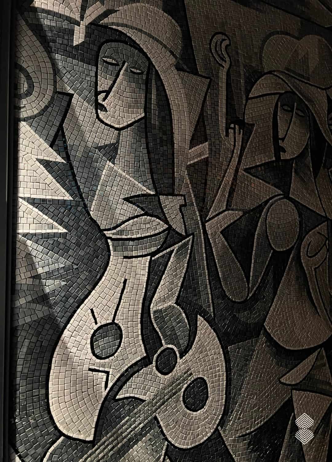 A tall cubist figures wall mosaic showing two musicians in fractured gray and black stone tesserae, installed on a cafe wall.