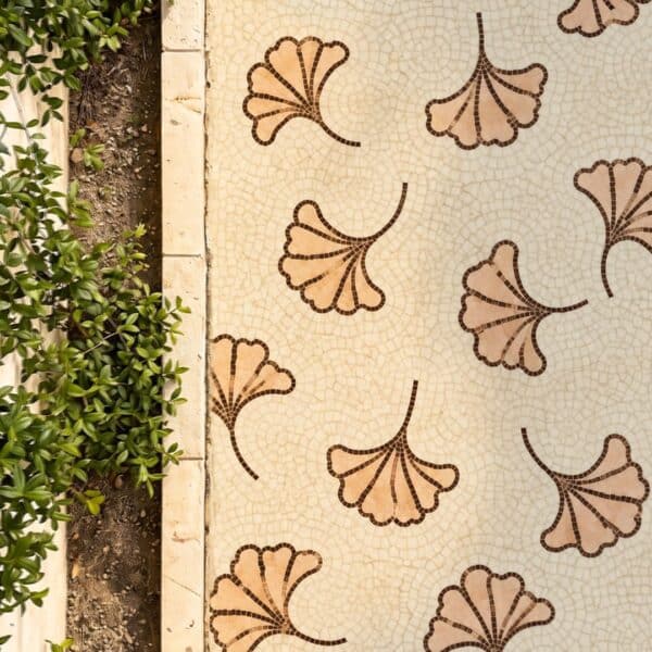 Ginkgo Leaf Mosaic Walkway A ginkgo leaf mosaic walkway shows scattered fan-shaped ginkgo leaves in tan and peach stone tesserae set into a cracked cream path beside a planted garden border.