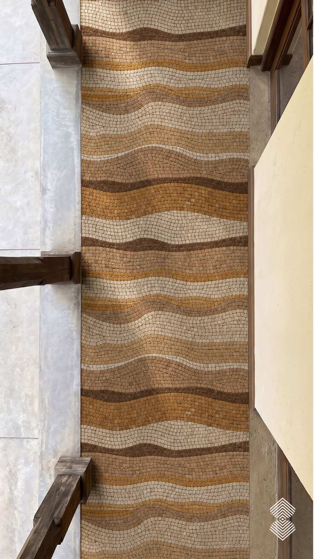 A long wave mosaic walkway panel of amber, gold and cream tesserae installed in a narrow entry corridor between two columns.