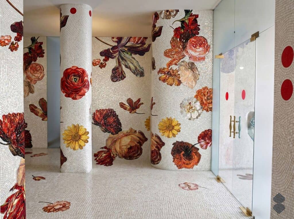 A grand interior entryway and room with mosaic tiles covering the floor, rounded pillars, and curved walls with vibrant red, orange, and yellow floral designs.