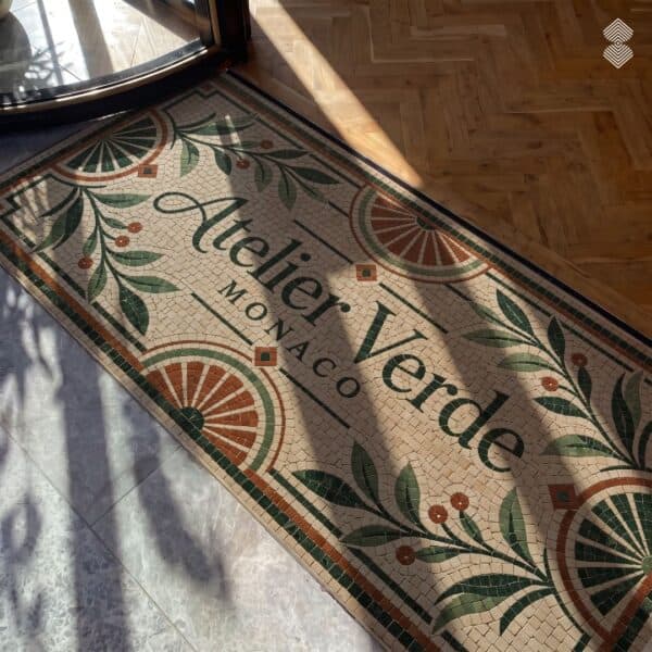A tumbled stone entrance logo mosaic mat with cursive script lettering framed by a green and terracotta leaf border at a doorway threshold.