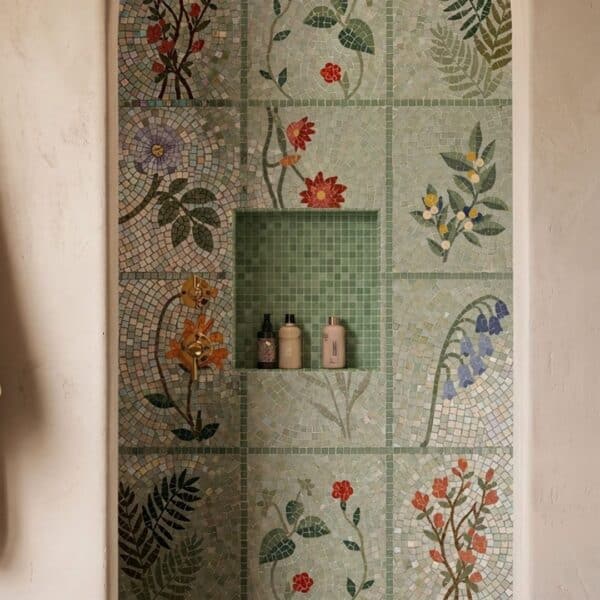 A grid of mosaic tile squares each depicting a different flower or fern on a pale green ground, installed in a shower niche.