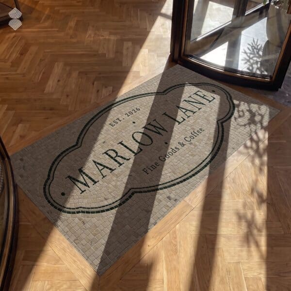 A custom logo floor mosaic in tumbled stone set into an entryway floor with serif lettering inside a scalloped oval frame.