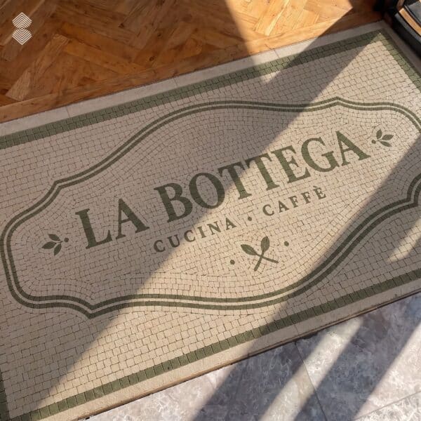 Cafe logo mosaic mat in tumbled stone set into a doorway floor with a serif wordmark inside a green-bordered cartouche.