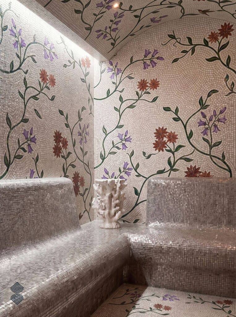 A tiled steam room featuring silver mosaic benches and white tile walls adorned with winding green vines and delicate purple and orange flowers.