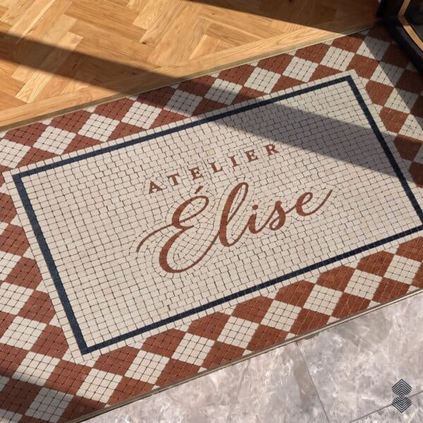 A script logo entryway mosaic set into a stone floor at a doorway, framed by a terracotta and cream diamond border.