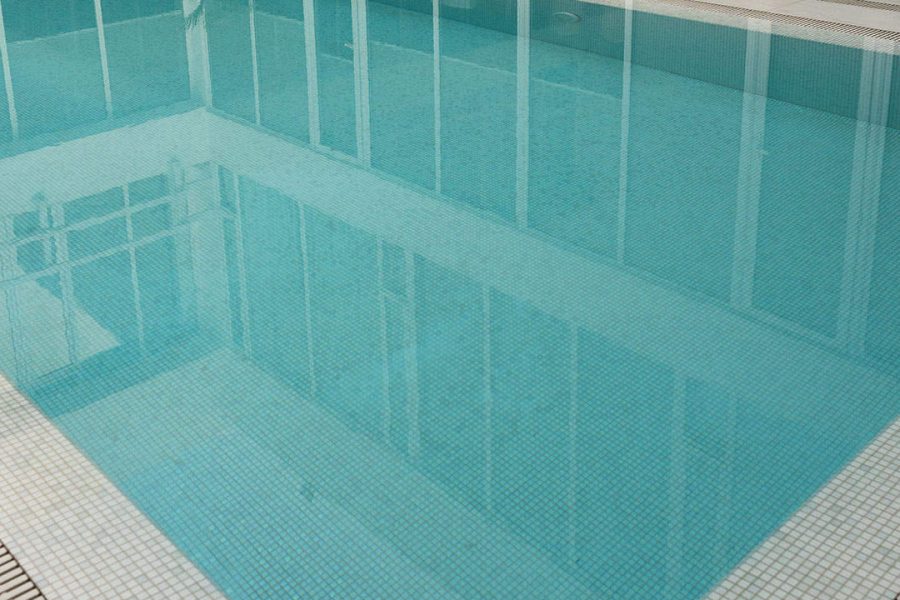 Clear blue-tiled swimming pool reflecting surrounding glass windows and white tiled edges.