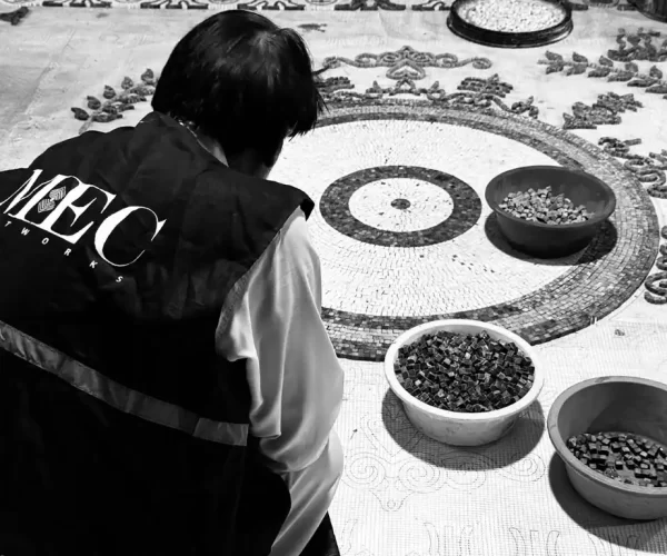 MEC artisan arranging mosaic tiles on circular pattern floor, surrounded by bowls of glass pieces.