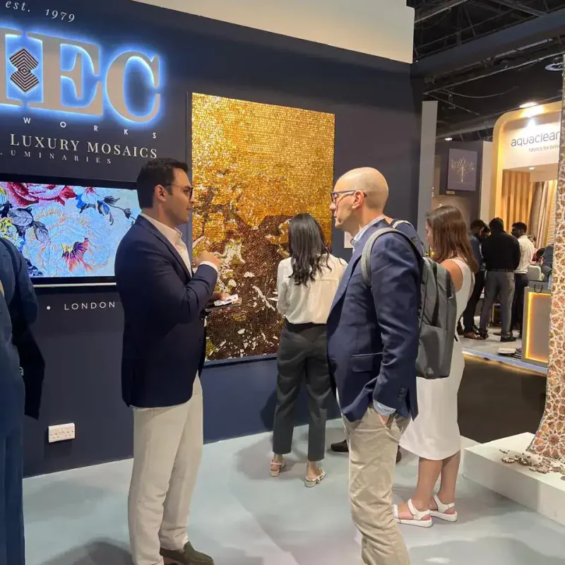 Two men converse at MEC booth near golden mosaic artwork during a luxury design exhibition.