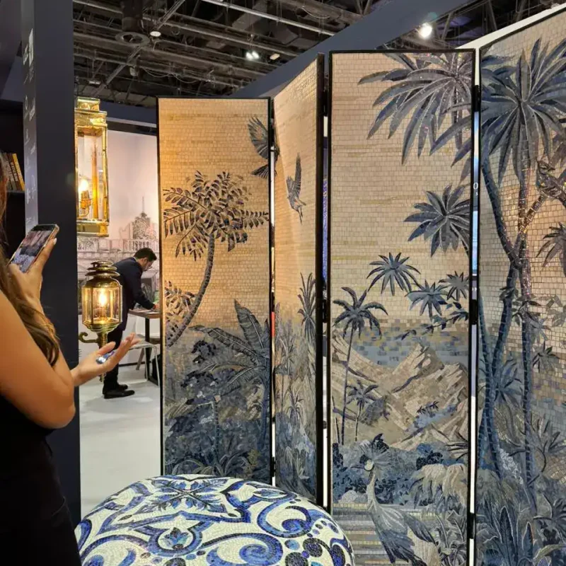 Woman photographing a mosaic jungle-themed folding screen beside a blue-patterned ottoman at an exhibit.