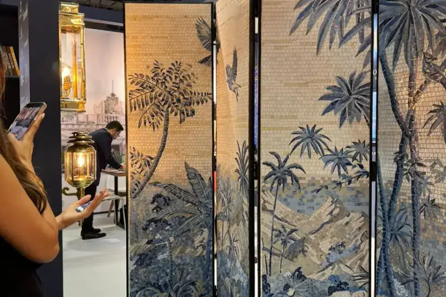 Woman photographing a mosaic jungle-themed folding screen beside a blue-patterned ottoman at an exhibit.