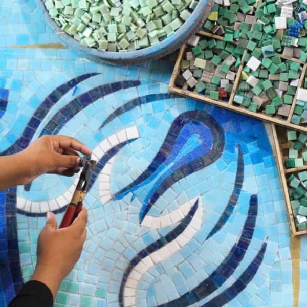 Artist working on blue mosaic using nippers and colored tiles for a wave-inspired design.
